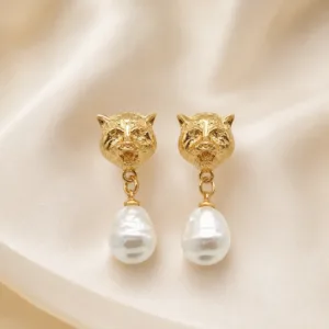 Brincos dourados com cabeças felinas e pérolas sobre cetim bege claro