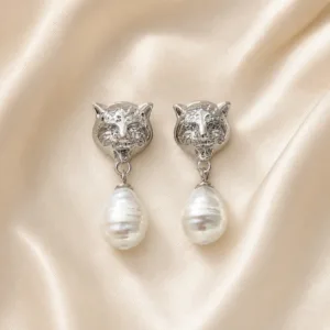 Brincos prateados com cabeças felinas e pérolas sobre cetim bege claro