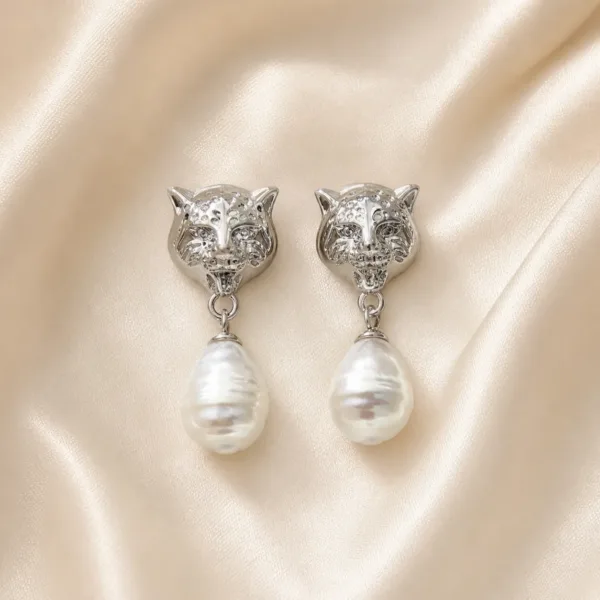 Brincos prateados com cabeças felinas e pérolas sobre cetim bege claro